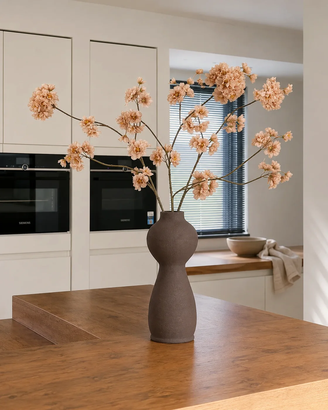 Nela-kunstboeket met cappuccinokleurige bloemen op luchtige takken in een bruine vaas op een houten kookeiland.