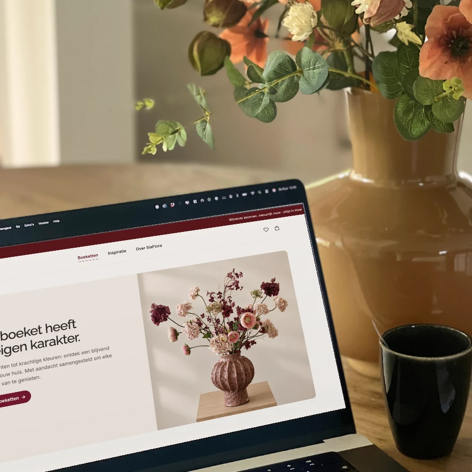 Werkplek van SiaFlora met de boekettenpagina op een laptop, een kop koffie en terracottakleurige bloemen in een okergele vaas.