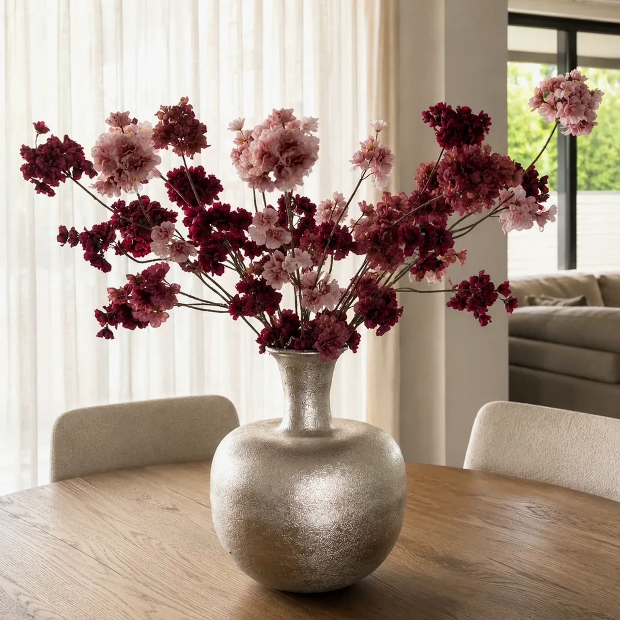 Zoya-boeket met roze en bordeauxrode bloesemtakken in een zilverkleurige vaas op een houten eettafel.