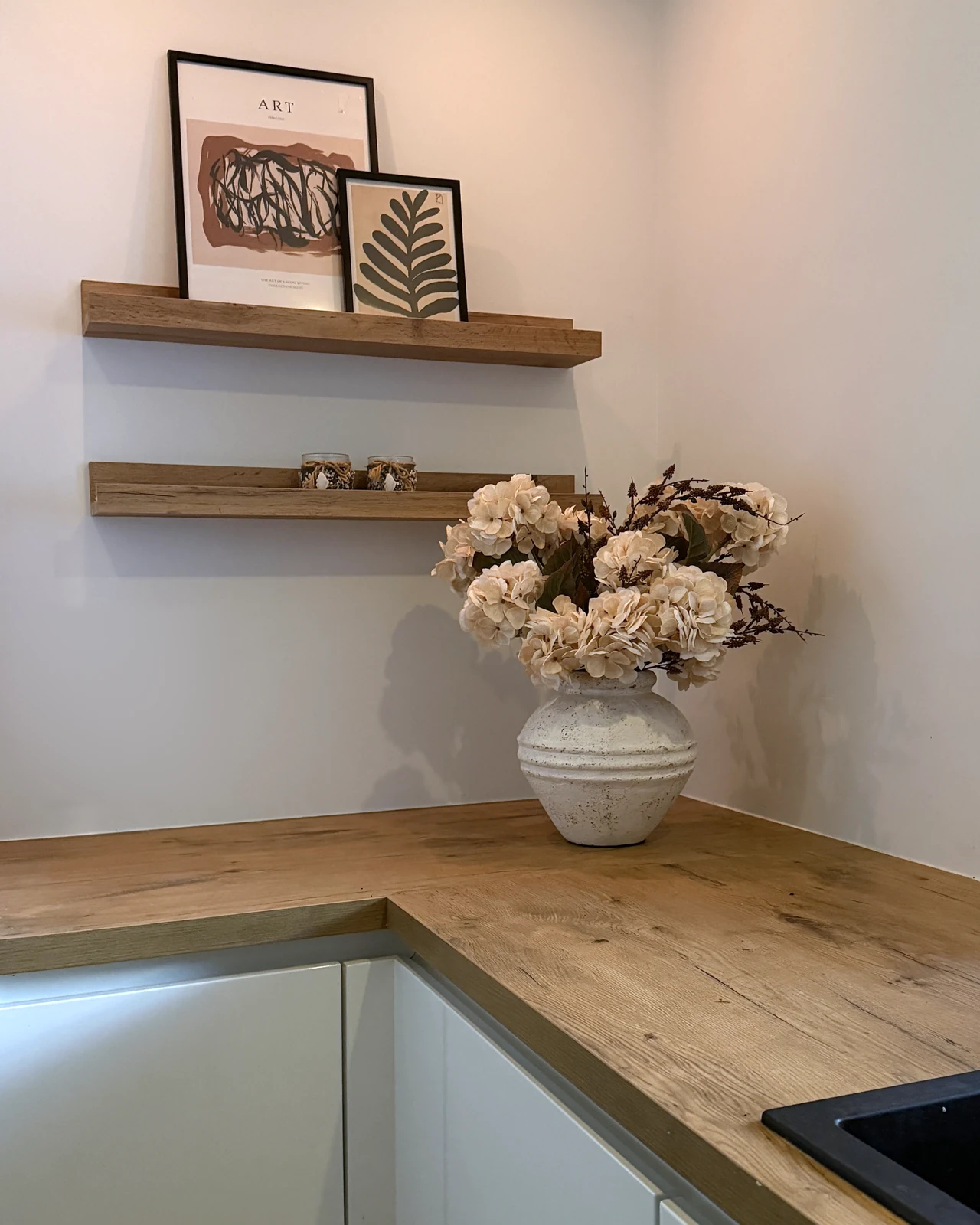 Odette, een crèmekleurig kunstboeket in een lichte vaas op een houten hoekaanrecht onder wandplanken.