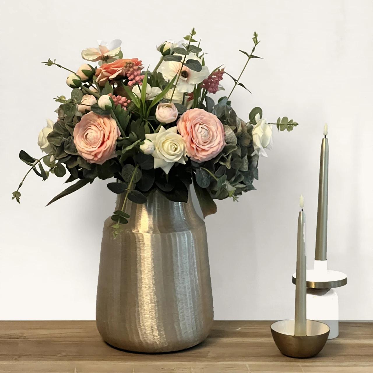 Nola, een kunstboeket met perzikkleurige en crèmekleurige bloemen in een goudkleurige vaas op een houten tafel naast twee kaarsen.