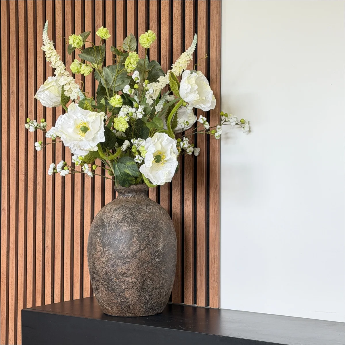 Witte en groene kunstbloemen in een donkere vaas op een zwart dressoir voor een houten lattenwand.