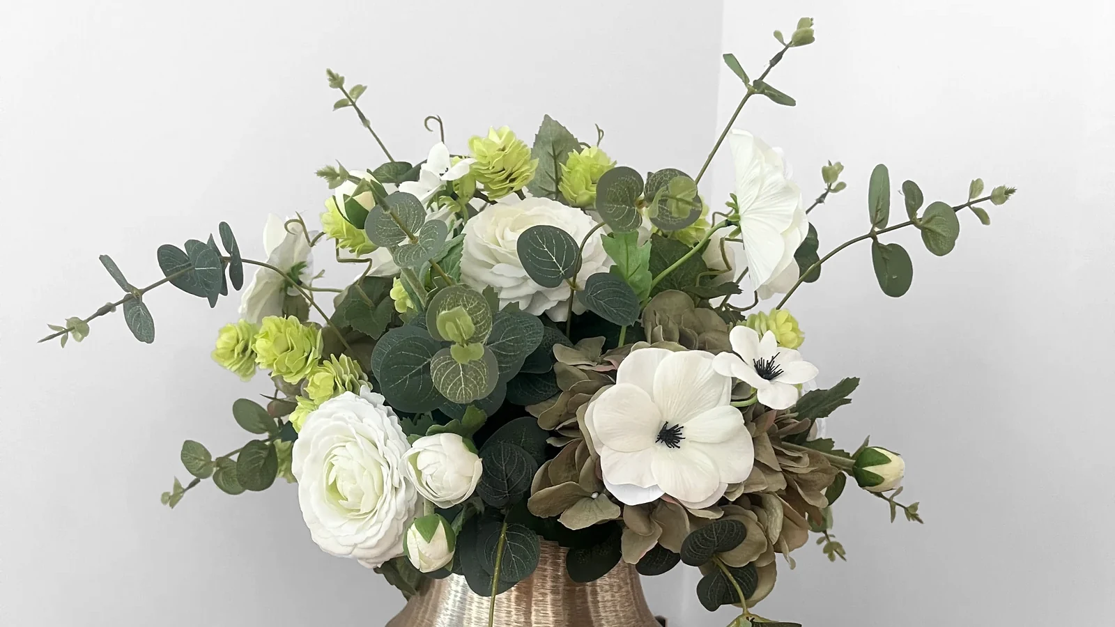 Jetta, een kunstboeket met witte en limegroene bloemen, taupekleurige hortensia en groen blad in een goudkleurige vaas.