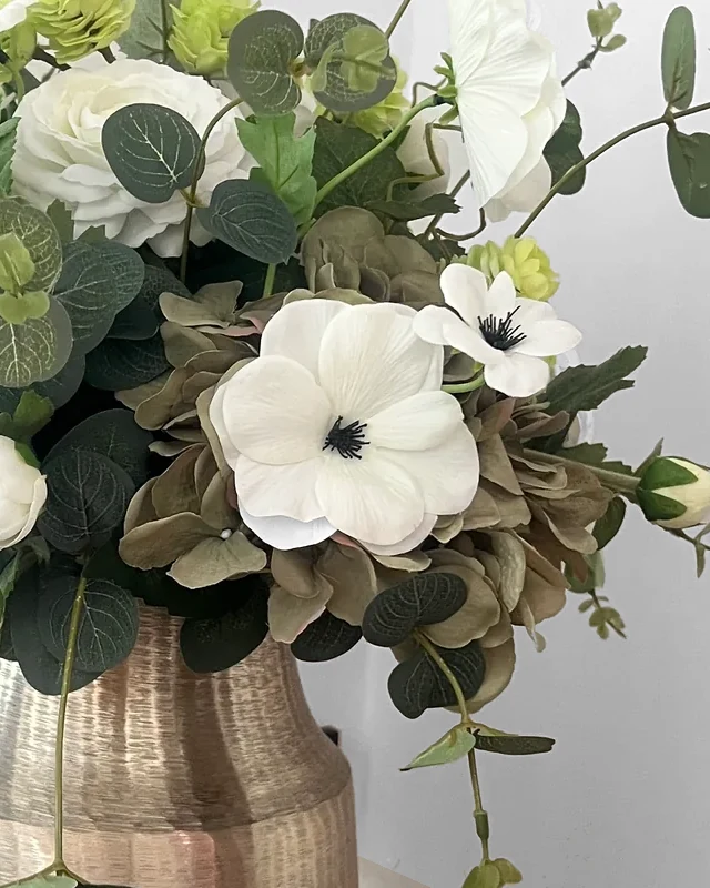 Detail van witte kunstbloemen met donkere harten tussen taupekleurige hortensia en groene bladeren van Jetta.
